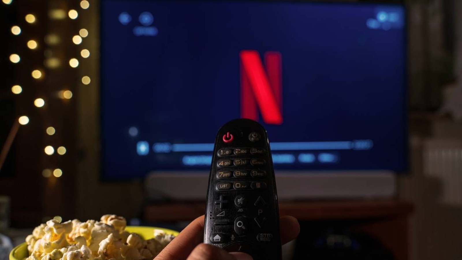 Mão segura controle remoto apontado para TV exibindo o logo Netflix, com pipoca