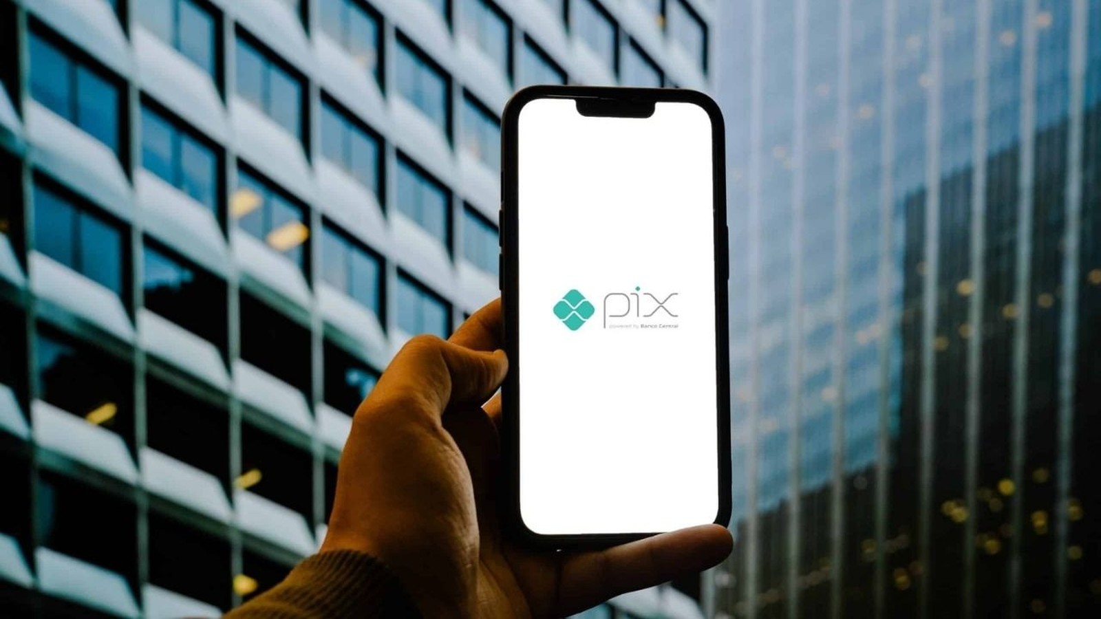 Mão segura smartphone com logo PIX na tela. Edifício moderno com janelas ao fundo