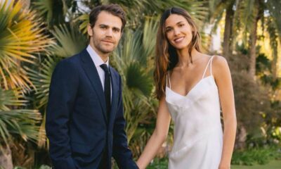 Paul Wesley de terno e Natalie Kuckenburg de vestido branco sorriem em jardim tropical