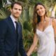 Paul Wesley de terno e Natalie Kuckenburg de vestido branco sorriem em jardim tropical