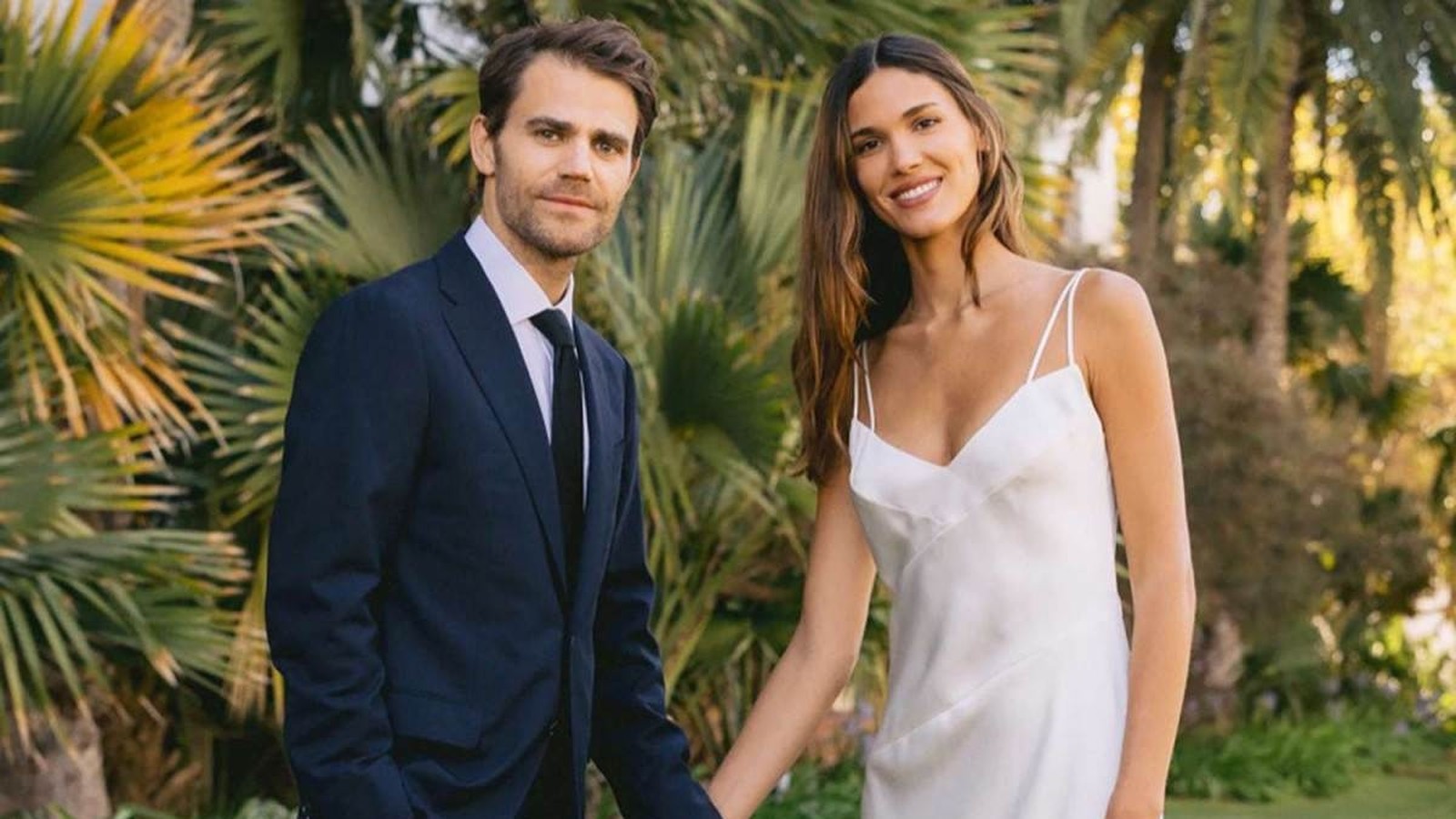 Paul Wesley de terno e Natalie Kuckenburg de vestido branco sorriem em jardim tropical