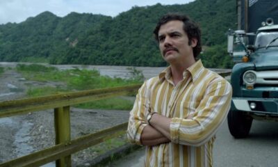 Wagner Moura (Pablo Escobar) de camisa listrada, braços cruzados, com montanhas e rio ao fundo