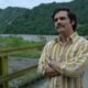 Wagner Moura (Pablo Escobar) de camisa listrada, braços cruzados, com montanhas e rio ao fundo