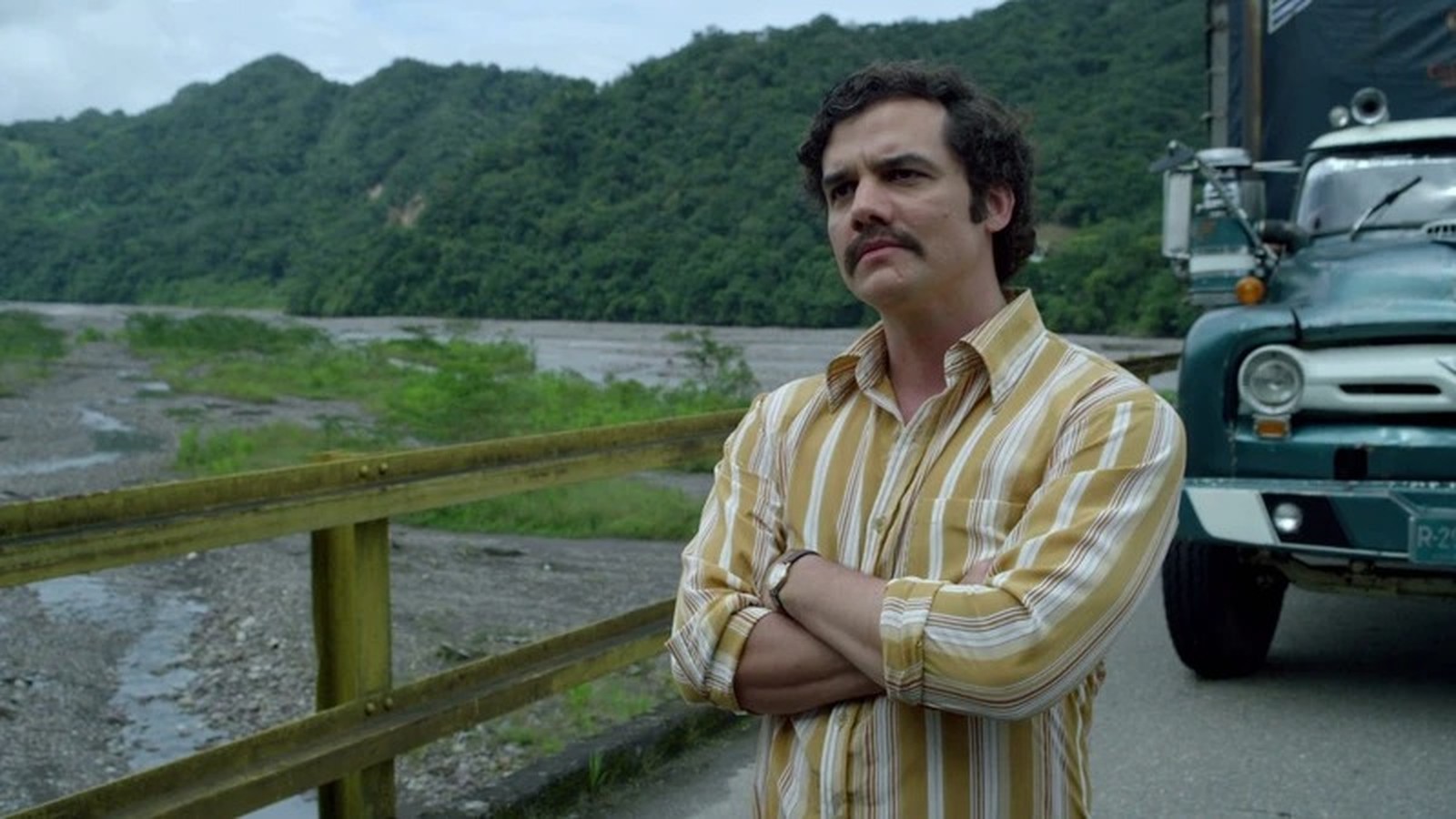 Wagner Moura (Pablo Escobar) de camisa listrada, braços cruzados, com montanhas e rio ao fundo