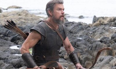 Matt Damon como Odisseu, barba, armadura, arco e flechas, em cenário rochoso de praia