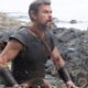 Matt Damon como Odisseu, barba, armadura, arco e flechas, em cenário rochoso de praia