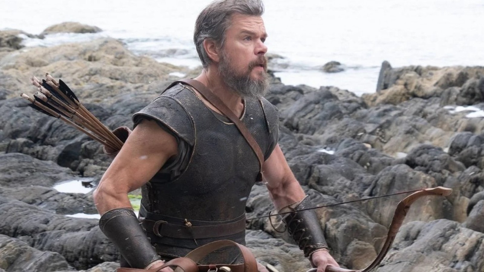 Matt Damon como Odisseu, barba, armadura, arco e flechas, em cenário rochoso de praia
