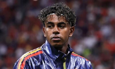 Close-up de Lamine Yamal, jovem com cabelo encaracolado e faixa branca, vestindo jaqueta esportiva azul