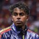Close-up de Lamine Yamal, jovem com cabelo encaracolado e faixa branca, vestindo jaqueta esportiva azul