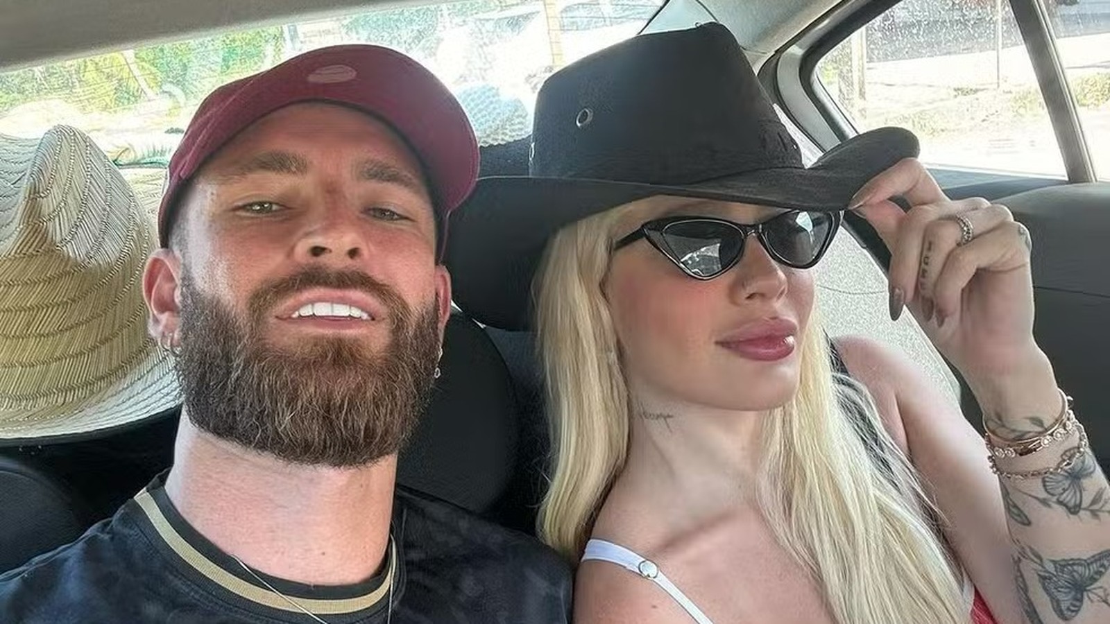 Selfie de casal no carro: homem de boné, mulher loira de chapéu e óculos escuros
