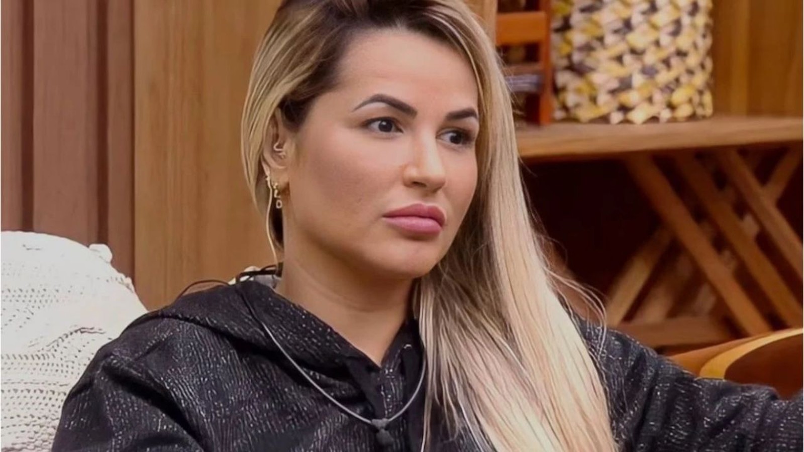 Influenciadora Deolane Bezerra com cabelo loiro, olhar pensativo, vestindo blusa preta, brincos em ambiente interno
