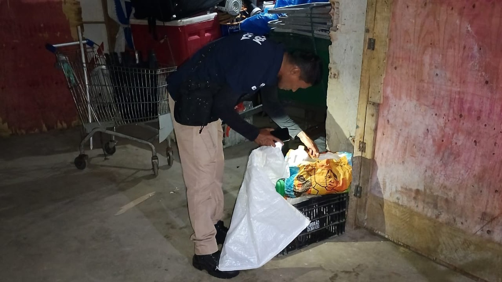 Homem em depósito inspeciona mercadorias apreendidas com saco e lanterna do celular