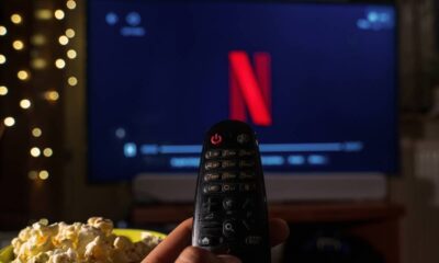 Mão segura controle remoto com tela de TV exibindo logo Netflix, pipoca e luzes ao fundo