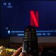 Mão segura controle remoto com tela de TV exibindo logo Netflix, pipoca e luzes ao fundo
