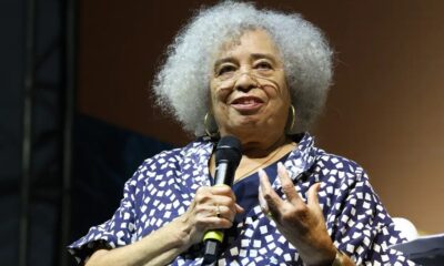 Angela Davis sorri, cabelo grisalho e óculos, segurando microfone e gesticulando