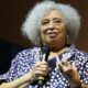 Angela Davis sorri, cabelo grisalho e óculos, segurando microfone e gesticulando
