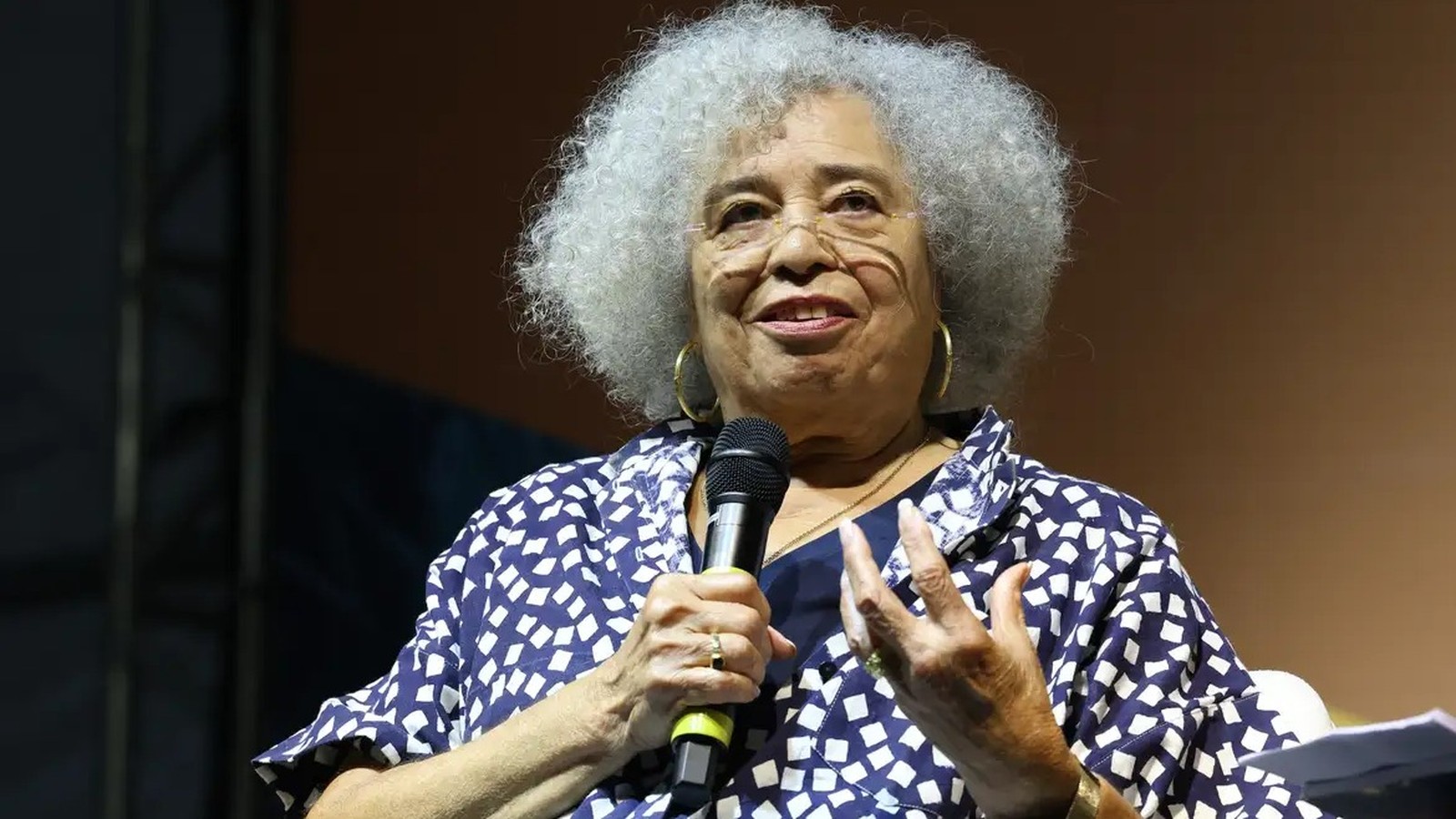 Angela Davis sorri, cabelo grisalho e óculos, segurando microfone e gesticulando