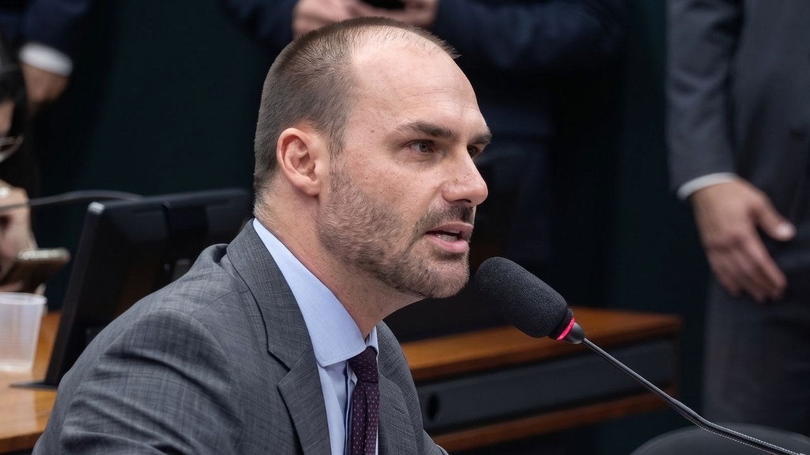 Eduardo Bolsonaro, homem branco com barba, terno cinza, falando em um microfone