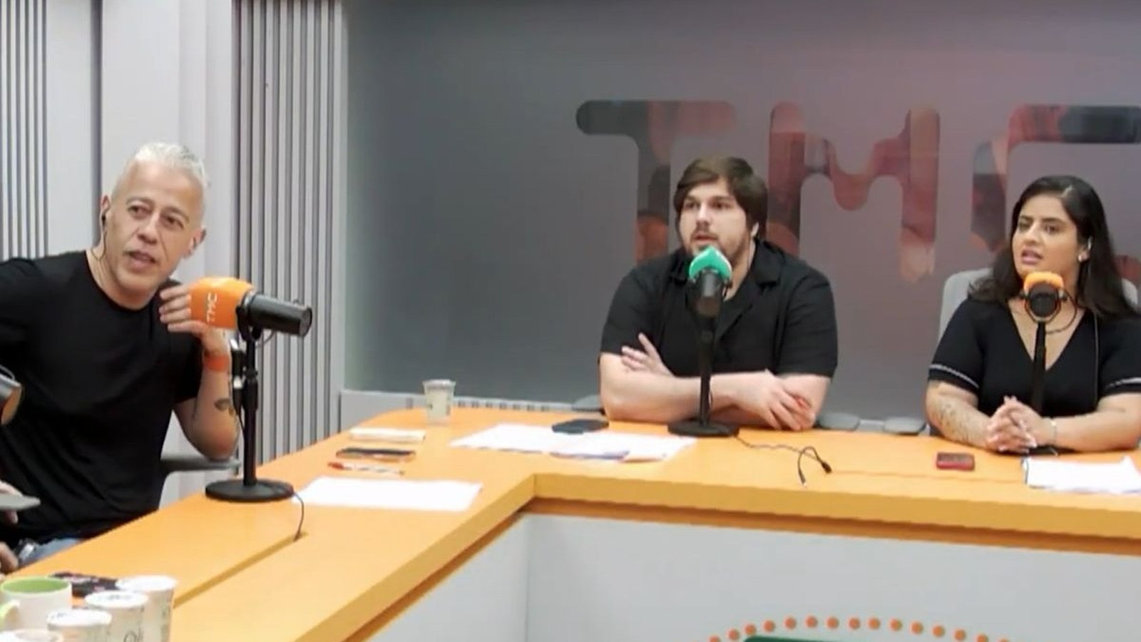 Alexandre Frota e dois apresentadores em estúdio de podcast, com microfones e mesa laranja