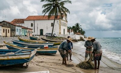 Pescadores na praia com rede, barcos, casas coloridas e igreja colonial.