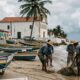 Pescadores na praia com rede, barcos, casas coloridas e igreja colonial.