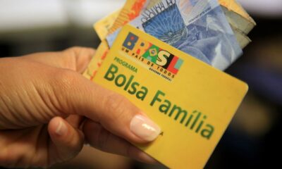 Mão segura cartão amarelo do Bolsa Família com notas de Real, mostrando o logo do programa.