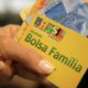 Mão segura cartão amarelo do Bolsa Família com notas de Real, mostrando o logo do programa.