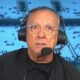 Galvão Bueno com óculos e fone de ouvido com microfone, em cabine de transmissão de estádio, com 'neo química' no fundo.
