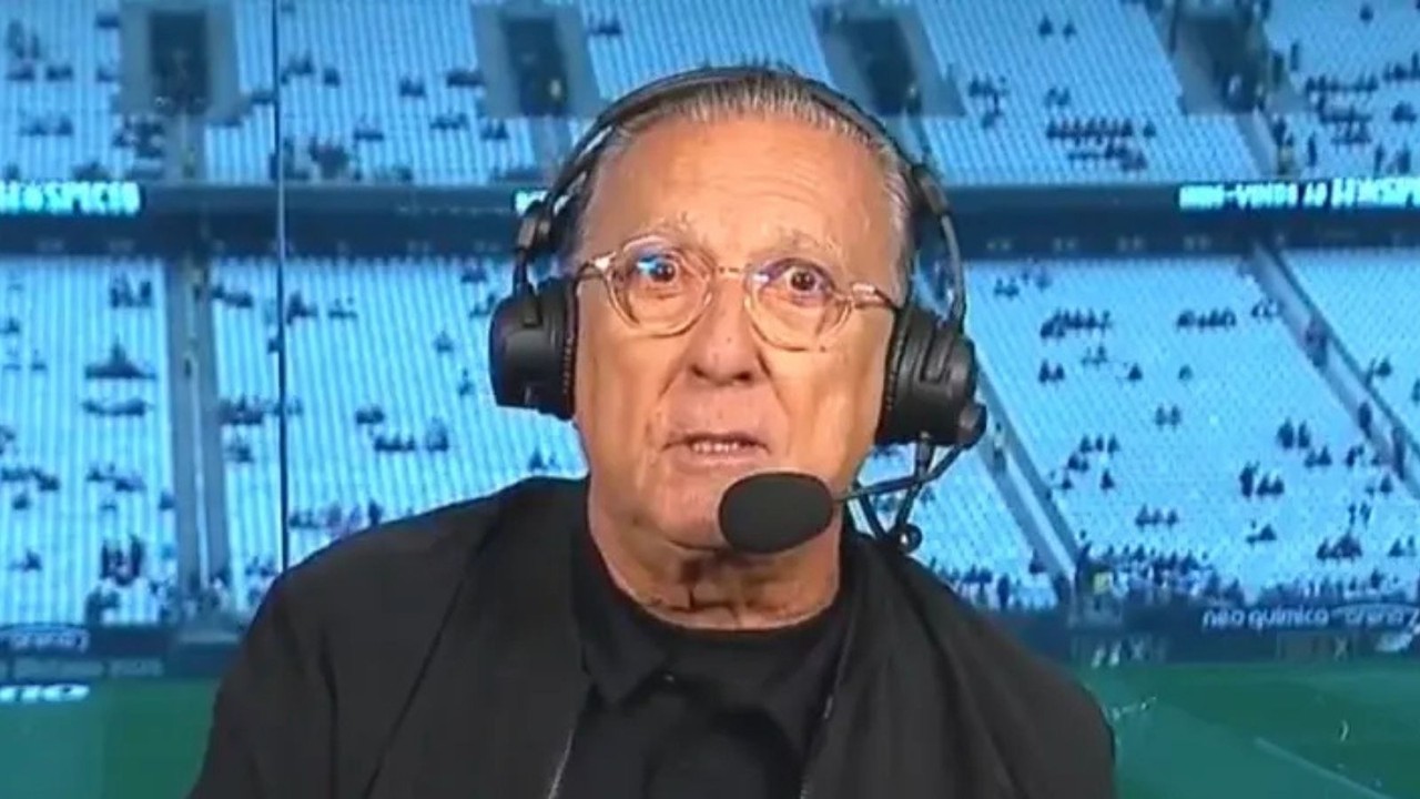 Galvão Bueno com óculos e fone de ouvido com microfone, em cabine de transmissão de estádio, com 'neo química' no fundo.