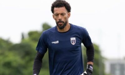 Goleiro de barba e cabelo cacheado, vestindo uniforme azul e luvas, em campo.