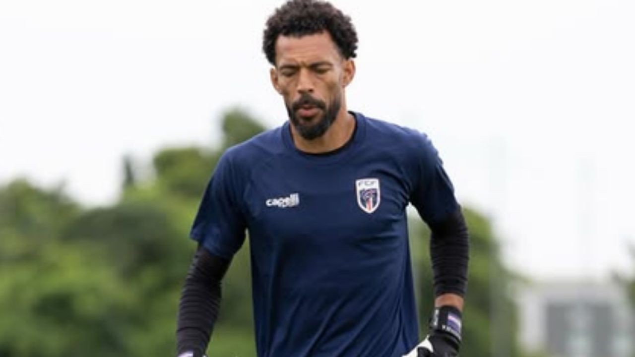 Goleiro de barba e cabelo cacheado, vestindo uniforme azul e luvas, em campo.