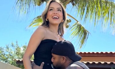 Homem de boné beija a barriga de mulher sorrindo sob céu azul e palmeiras