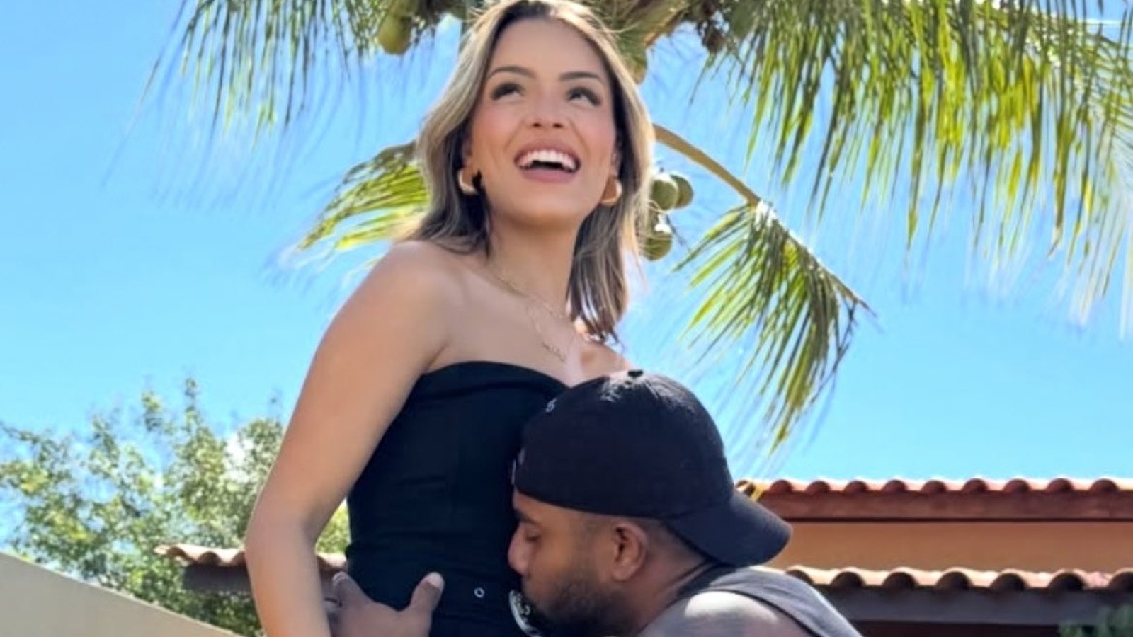 Homem de boné beija a barriga de mulher sorrindo sob céu azul e palmeiras