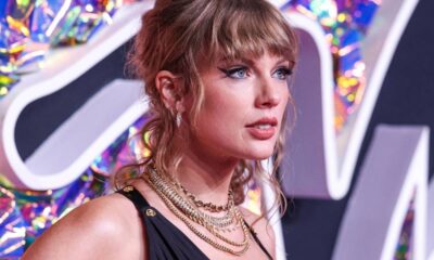Taylor Swift de perfil, cabelo loiro, olhos azuis, maquiagem, colares dourados, fundo colorido.