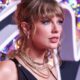 Taylor Swift de perfil, cabelo loiro, olhos azuis, maquiagem, colares dourados, fundo colorido.