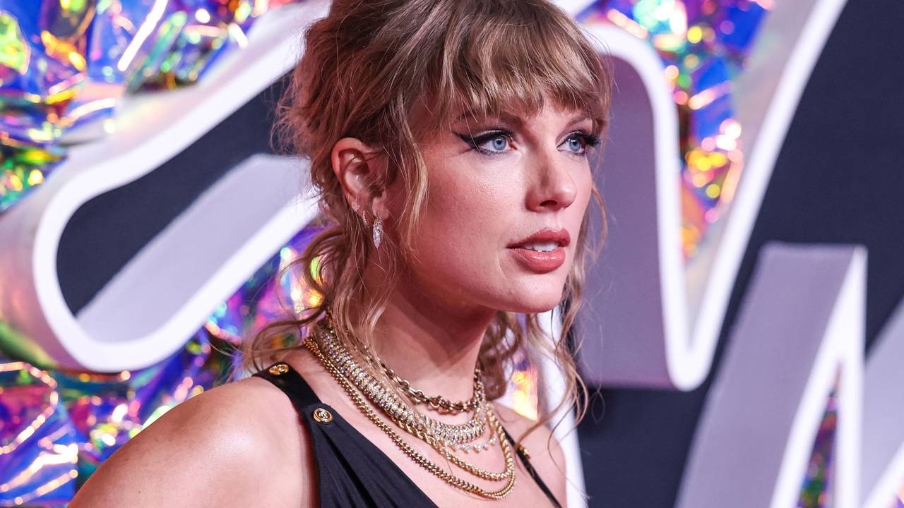 Taylor Swift de perfil, cabelo loiro, olhos azuis, maquiagem, colares dourados, fundo colorido.