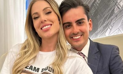 Casal sorrindo para a câmera, homem de terno e mulher de camiseta branca.