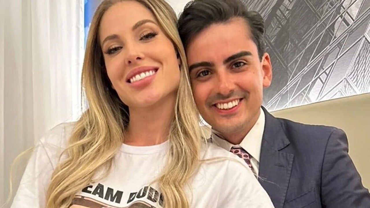 Casal sorrindo para a câmera, homem de terno e mulher de camiseta branca.
