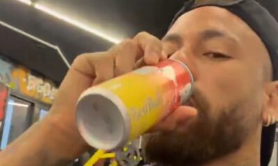 Homem com barba e boné preto bebendo de uma lata de Red Bull amarela e vermelha