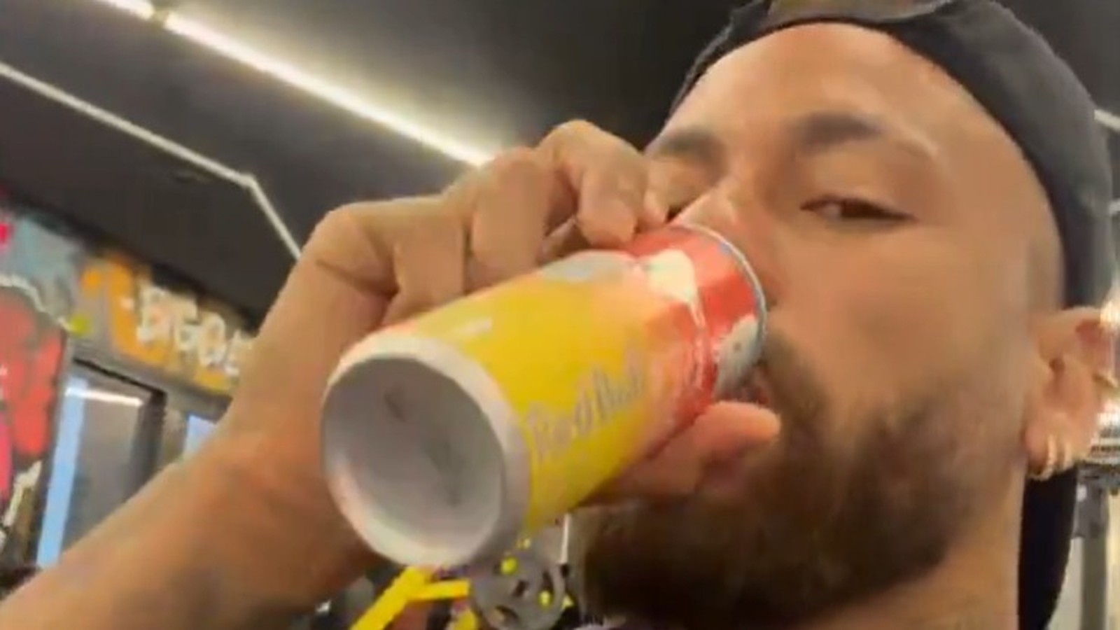 Homem com barba e boné preto bebendo de uma lata de Red Bull amarela e vermelha