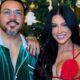 Belo e Rayane Figliuzzi sorrindo lado a lado, em frente a uma árvore de Natal decorada