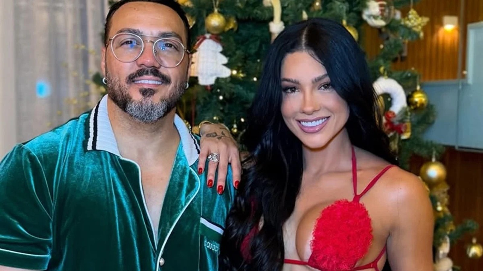 Belo e Rayane Figliuzzi sorrindo lado a lado, em frente a uma árvore de Natal decorada