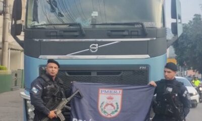Dois policiais militares com fuzil e bandeira PMERJ em frente a caminhão Volvo azul