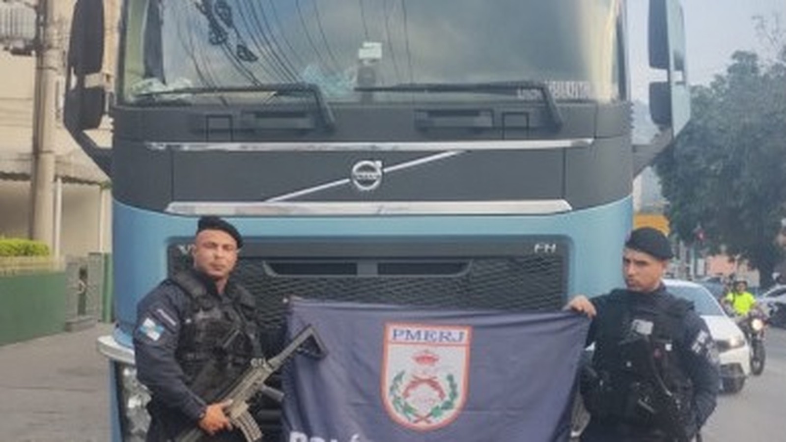 Dois policiais militares com fuzil e bandeira PMERJ em frente a caminhão Volvo azul