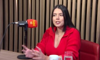 Fernanda Bande em estúdio de podcast, falando ao microfone da Rádio Tupi, usando suéter vermelho