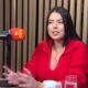 Fernanda Bande em estúdio de podcast, falando ao microfone da Rádio Tupi, usando suéter vermelho
