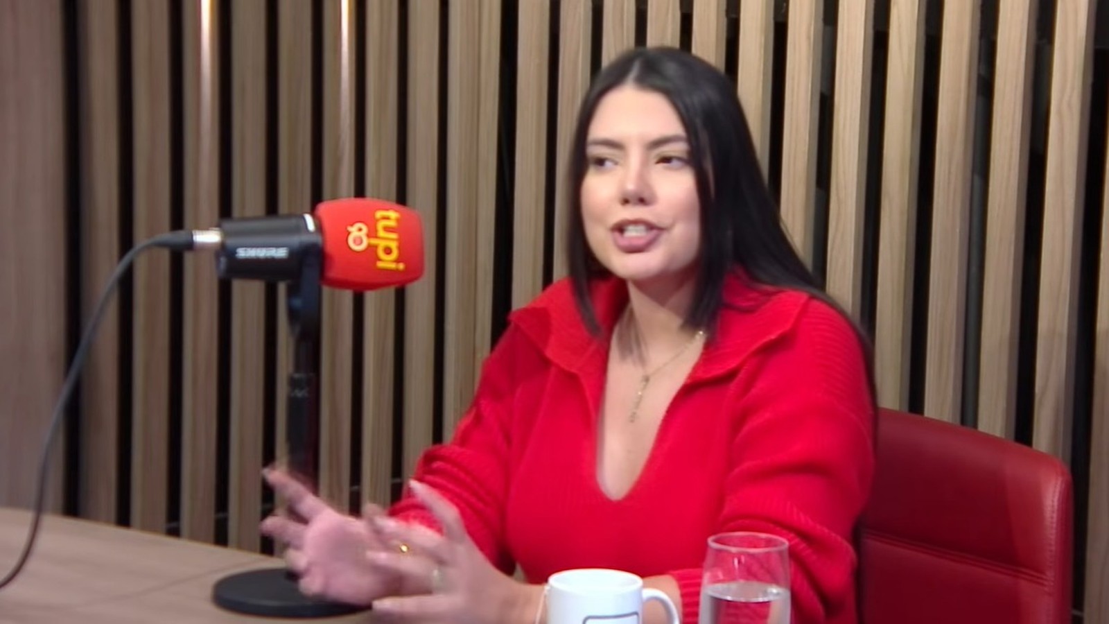 Fernanda Bande em estúdio de podcast, falando ao microfone da Rádio Tupi, usando suéter vermelho