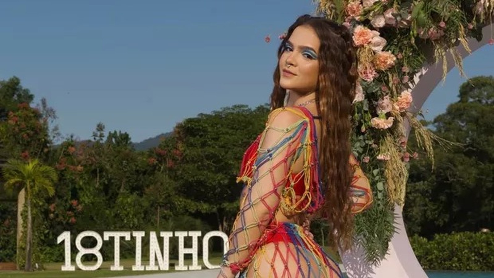 Mel Maia com roupa de crochê colorida, ao ar livre, com placa 18TINHO