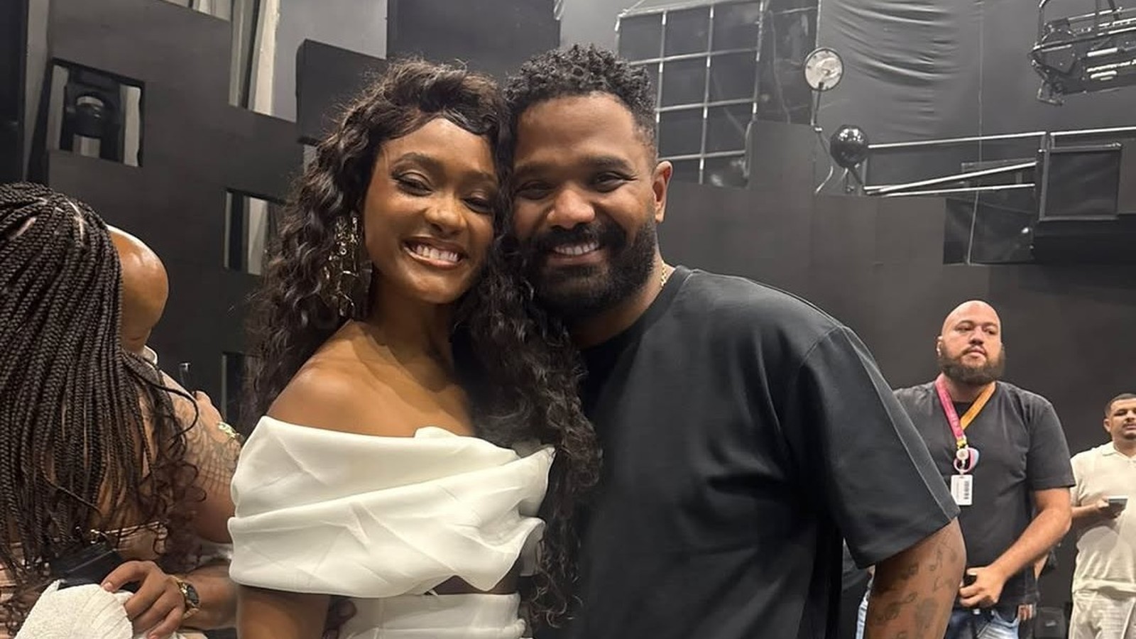 Erika Januza e Arlindinho sorrindo e abraçados em um estúdio com fundo escuro