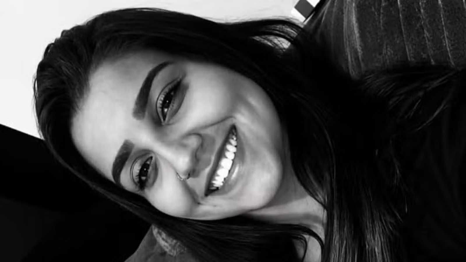 Close-up em preto e branco de mulher jovem sorrindo, com cabelo escuro e piercing no nariz